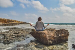 Eine junge Frau auf einem Fels in einer ruhigen Yogapose vor Meer mit Brandung.